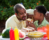 A son feeds his dad a hot dog.