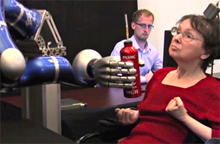 Photo of a woman controlling a robotic arm to grasp a bottle.