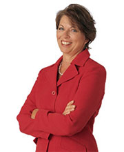 Brown haired woman with a short bob hair style wearing a red suit with white pearls with her hands crossed over her chest smiling.