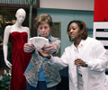 An older woman is checking her BMI as a health care professional in a white Dr.'s coat assists her. Both women are standing in front of a mannequin that is wearing a formal red dress.