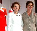 Mrs. Nancy Reagan in a white skirt suite and Mrs. Laura Bush in a gray skirt suit with a Red Dress Pin pose in front of several First Ladies red dresses.
