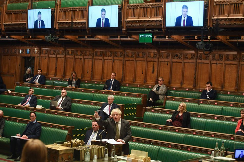 A photograph of MPs sat in the Chamber while Keir Starmer joins via videolink to question the Prime Minister during PMQs. Prime Minister Boris Johnson is pointing at the screen on which Mr Starmer appears.