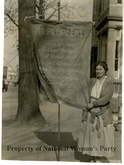 Mrs. Bartlet carring a "Mr President" banner. She stands in front of a building next to the banner holding its pole and the corner.