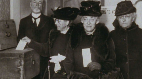 A black-and-white photo of women casting a vote by placing a slip of paper in a ballot box. It is clear from the antiquated dress that the photo is from the early twentieth century. | Video