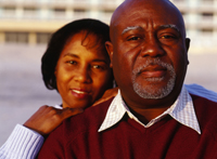 A man and a woman standing. The middle-aged man is in the foreground, looking into camera. The woman is standing behind him with her hands on his shoulder, smiling and also looking into the camera.
